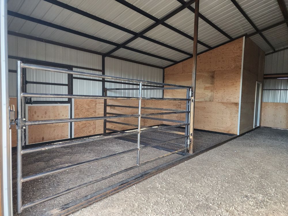 Interior open bay of a metal horse barn with a full-width steel gate and a plywood-walled tack room