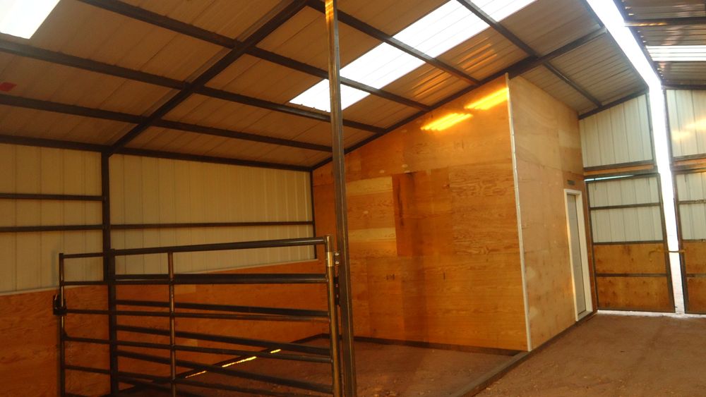 Center breezeway of a metal horse barn looking past a framed plywood tack room toward a full width steel gate