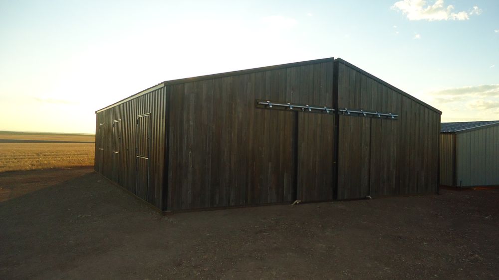 Three quarter view of a woodgrain metal horse barn at sunset showing the gable end sliding doors and the man doors on the tack room side